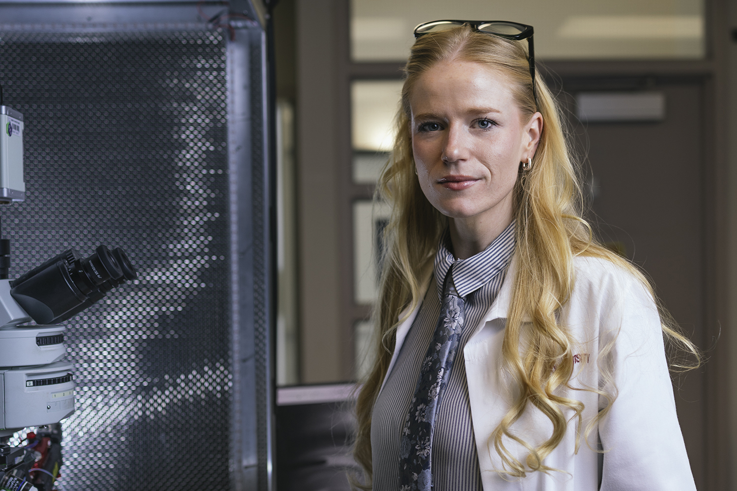 Scientist Annie Goettemoeller, with long blond hair, in a lab coat stands near a microscope, showcasing a research environment.