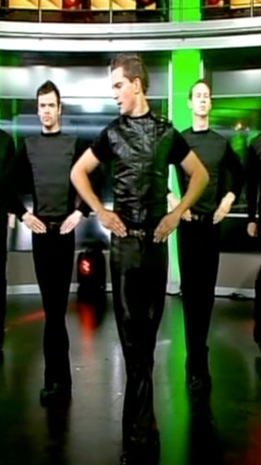 A dance performance featuring multiple male dancers in black attire, showcasing synchronized movements on a brightly lit stage.
