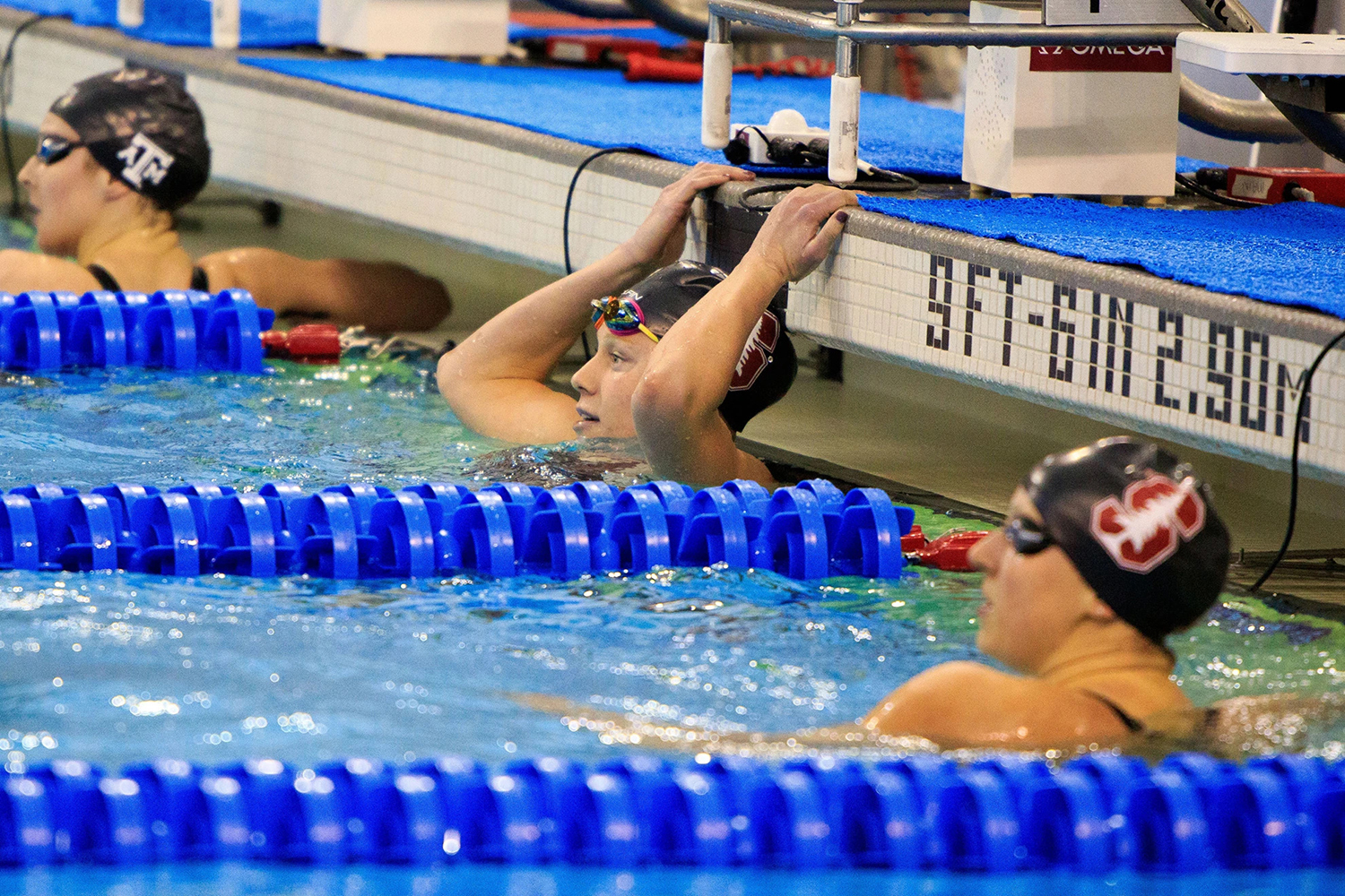 Ella Eastin and two other swimmers rest at the pool edge after a race, gripping the lane barrier among colorful lane dividers.