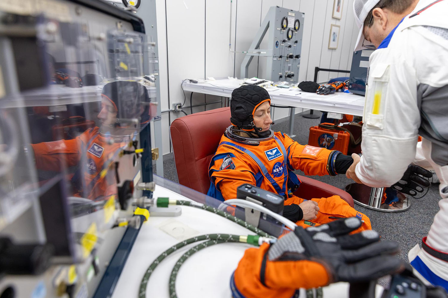 Astronaut in orange spacesuit sits in testing facility while technician assists.