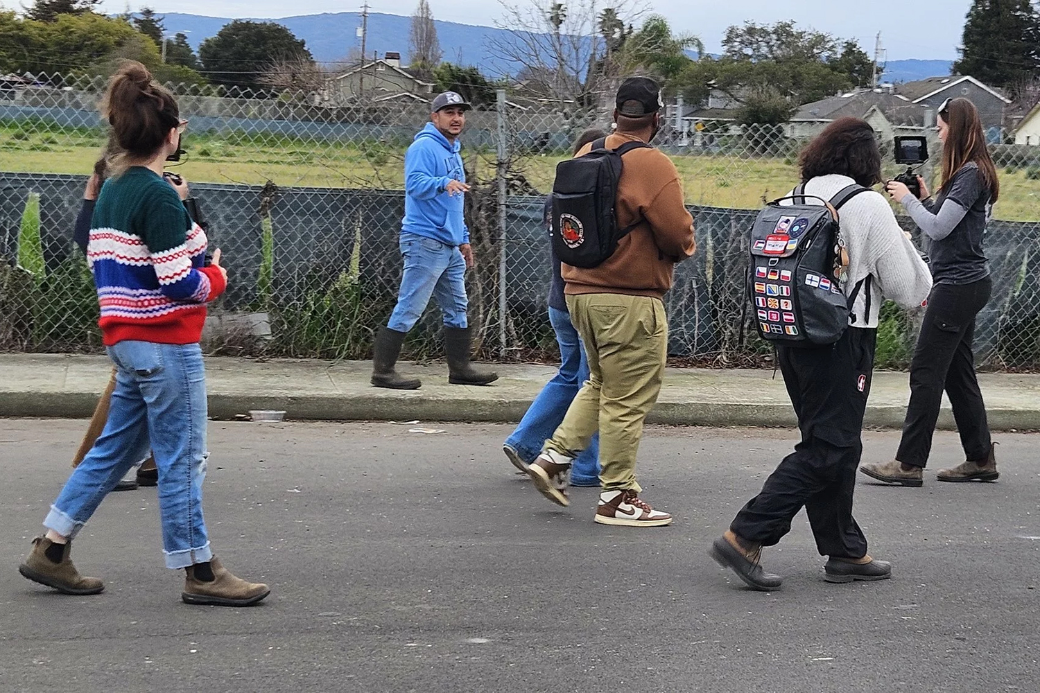 A group of five people walking along a street, wearing casual clothing, with one person filming a person in blue who is talking to the camera.