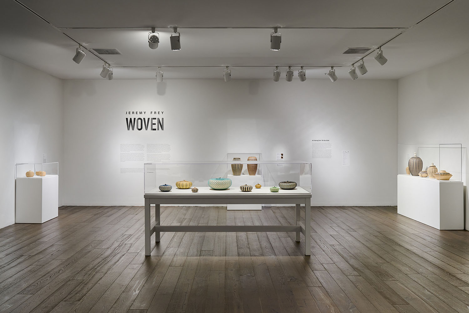 Gallery exhibition featuring woven baskets in display cases under track lighting with wall text reading Jeremy Frey: Woven.