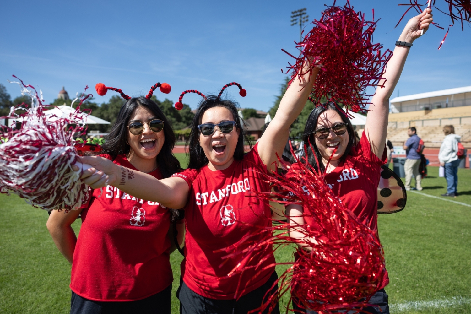 Three people in red Stanford shirts wave red pom-poms and celebrate at an outdoor event.
