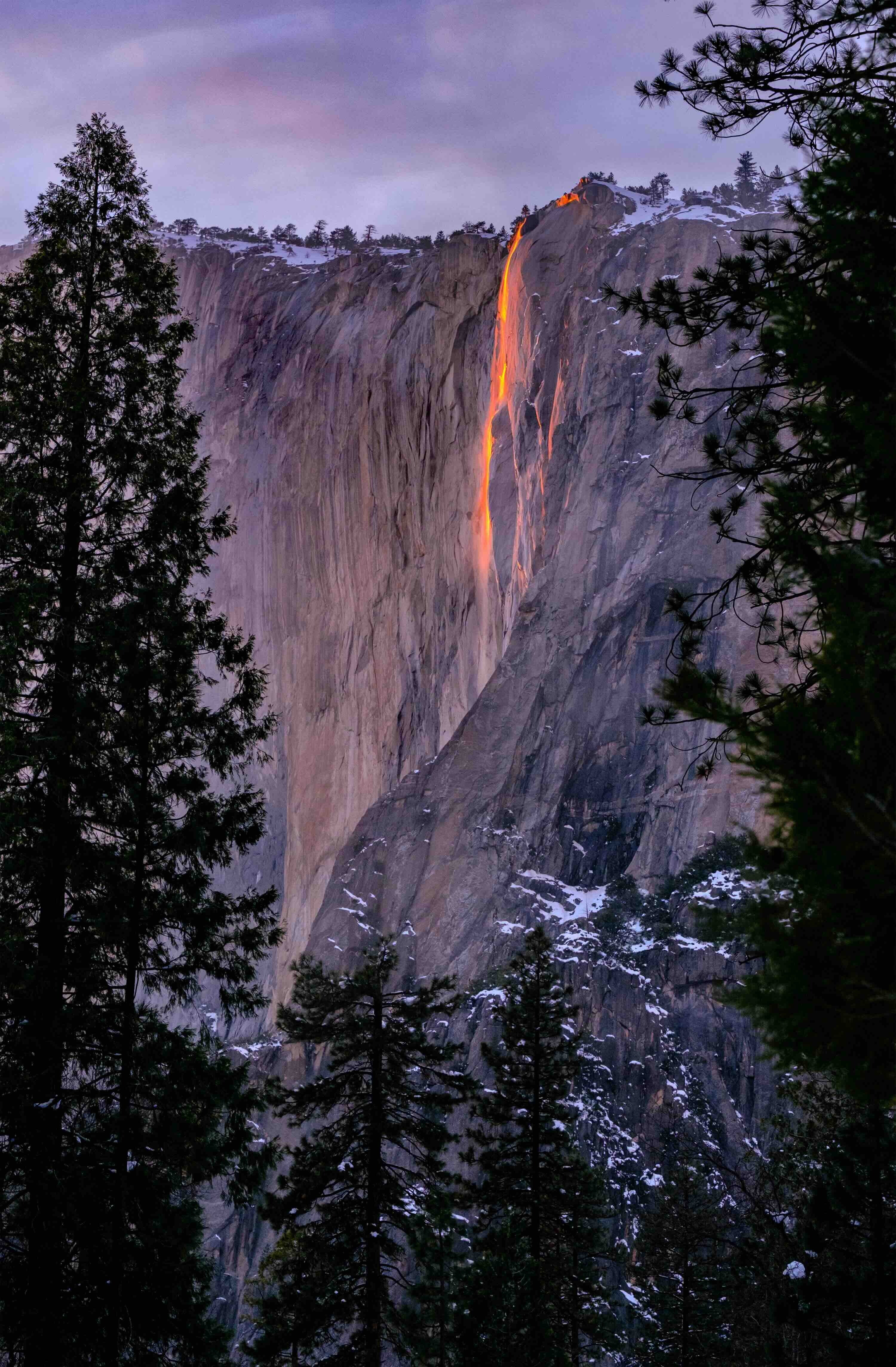 A glowing orange waterfall cascades down a rocky cliff, framed by dark green trees against a twilight sky.
