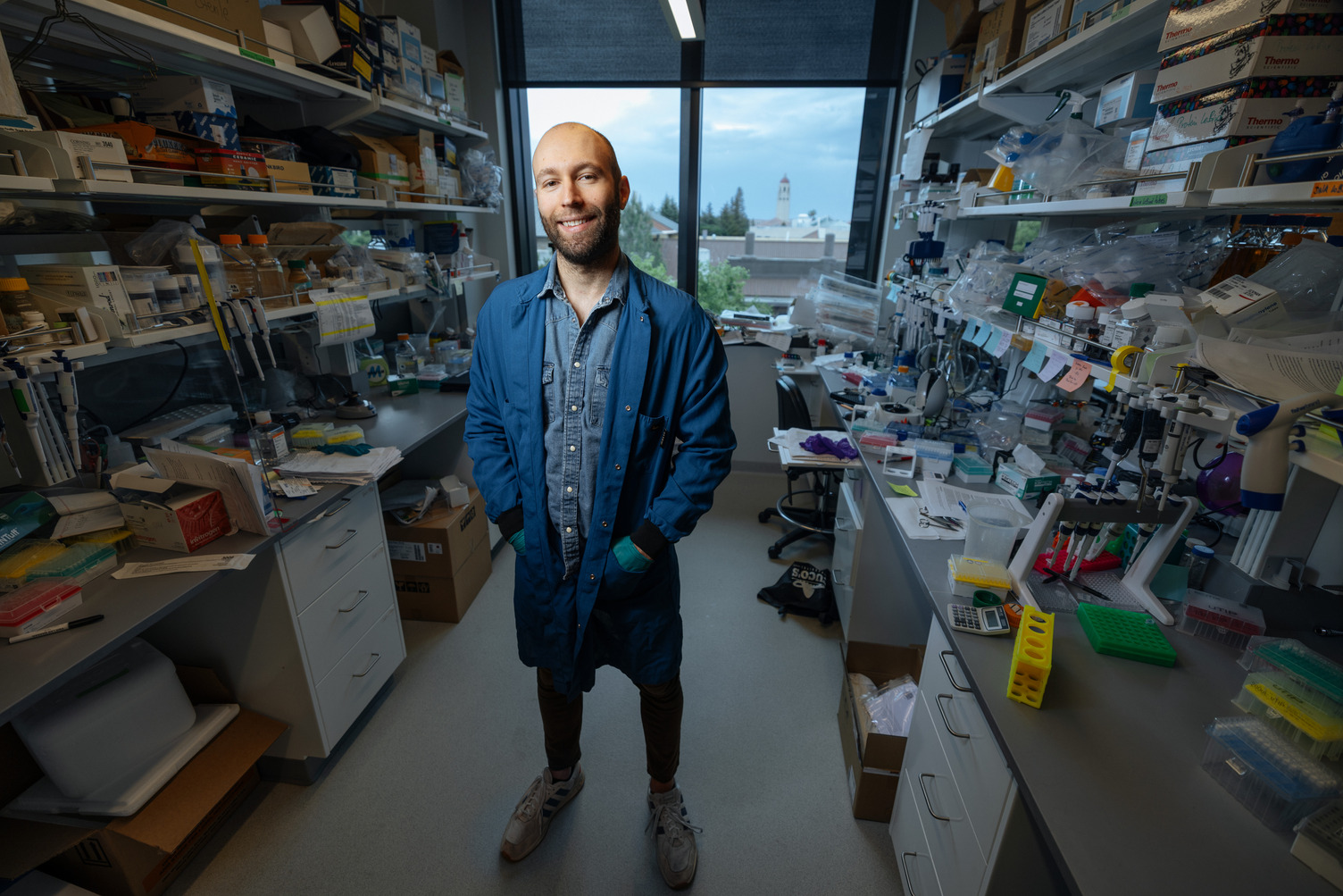 Blake Laham in a lab coat stands in a cluttered laboratory filled with scientific equipment and supplies, with windows showing an outdoor view.