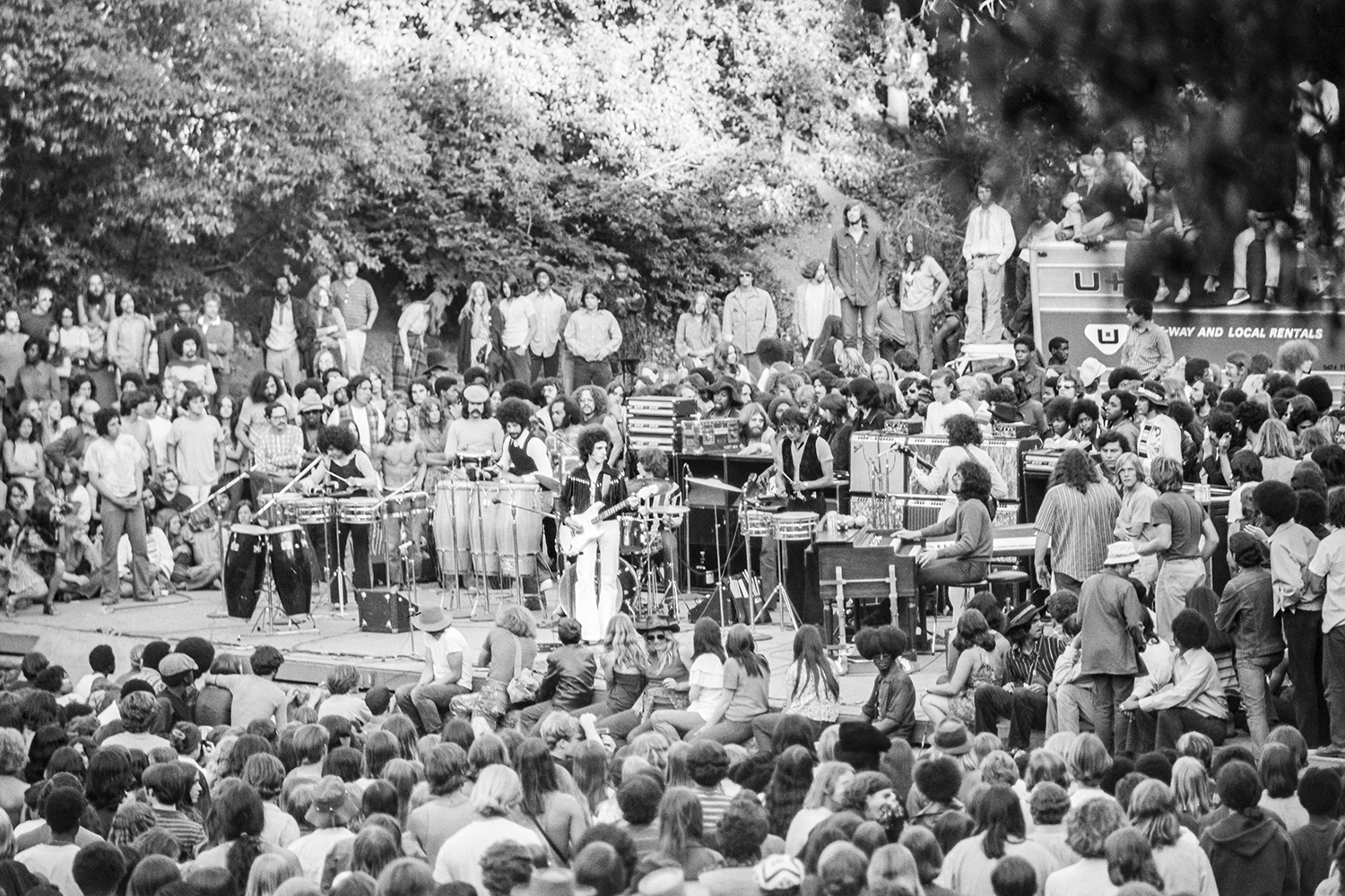 A large crowd gathers to watch Santana perform on stage with various instruments in an outdoor setting.