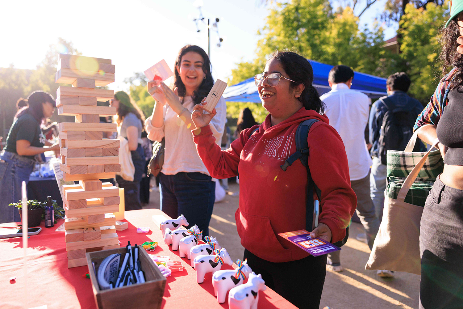 People explore a vibrant outdoor resource fair, with one woman holding a wooden block and pamphlet next to a tall stack of wooden stacking blocks and colorful toys.