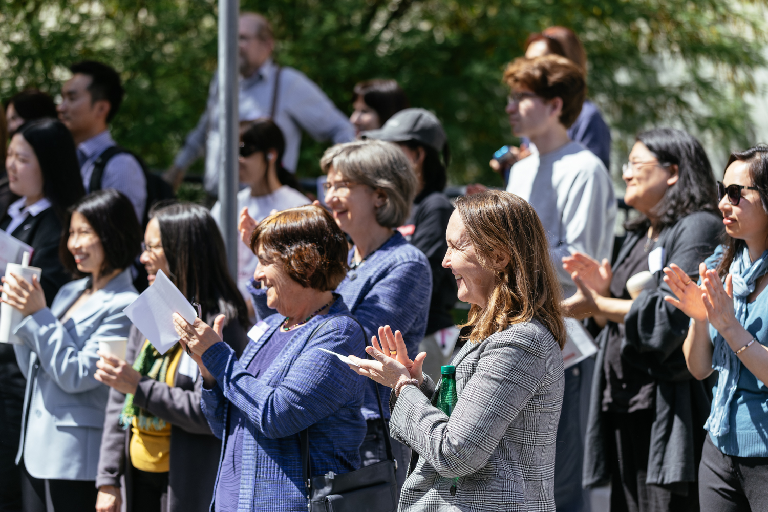 A diverse crowd of people applauds enthusiastically during an outdoor event, with many holding pamphlets or drinks.