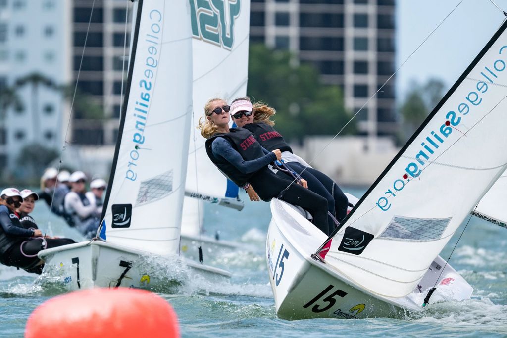Teams of sailors compete in a race, leaning into their boats to catch the wind, with a city skyline in the background.