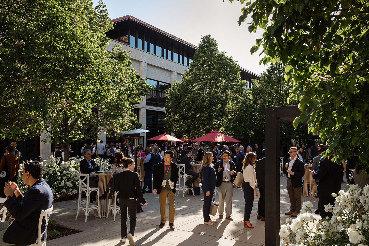 A lively outdoor gathering with people mingling near tables adorned with white flowers and shaded by large trees.