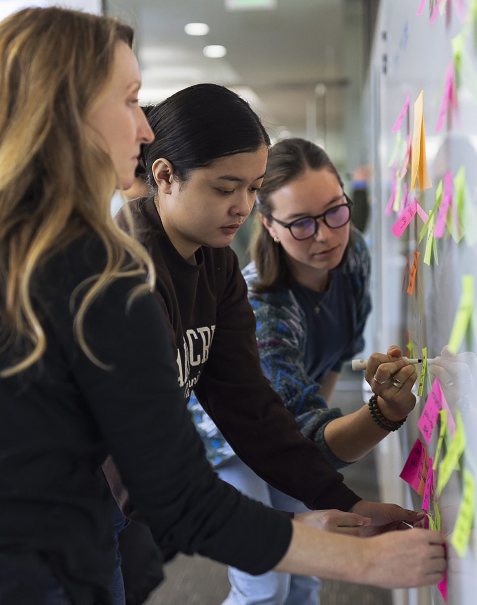 Three people work on a white board covered in different colored Post-it Notes. 