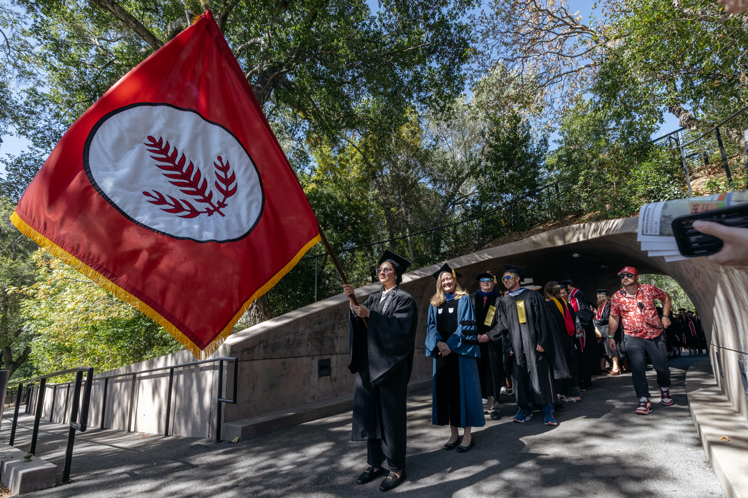 Graduates in caps and gowns walk under a tunnel, led by a student holding a flag featuring a laurel design.
