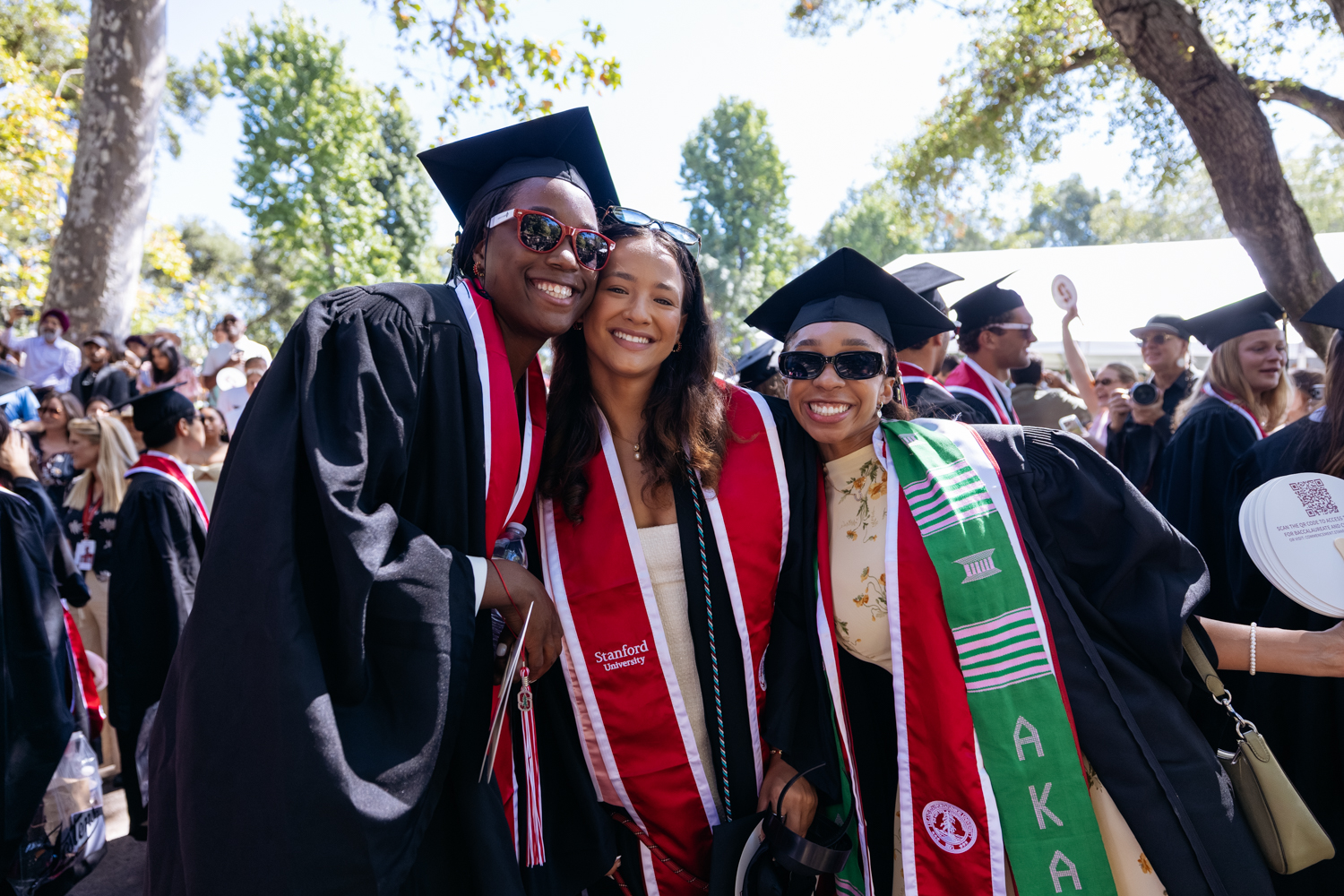 Graduates in caps and gowns pose together, wearing stoles, during a festive outdoor graduation ceremony.