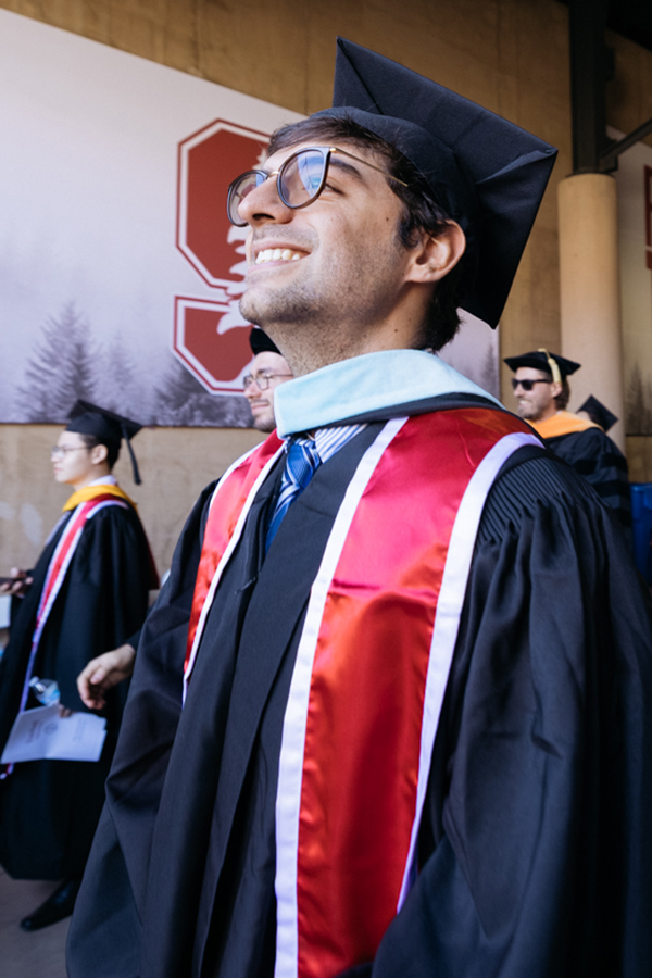Male graduate wearing glasses in cap and gown smiles while looking upward during entrance to stadium for commencement ceremony.