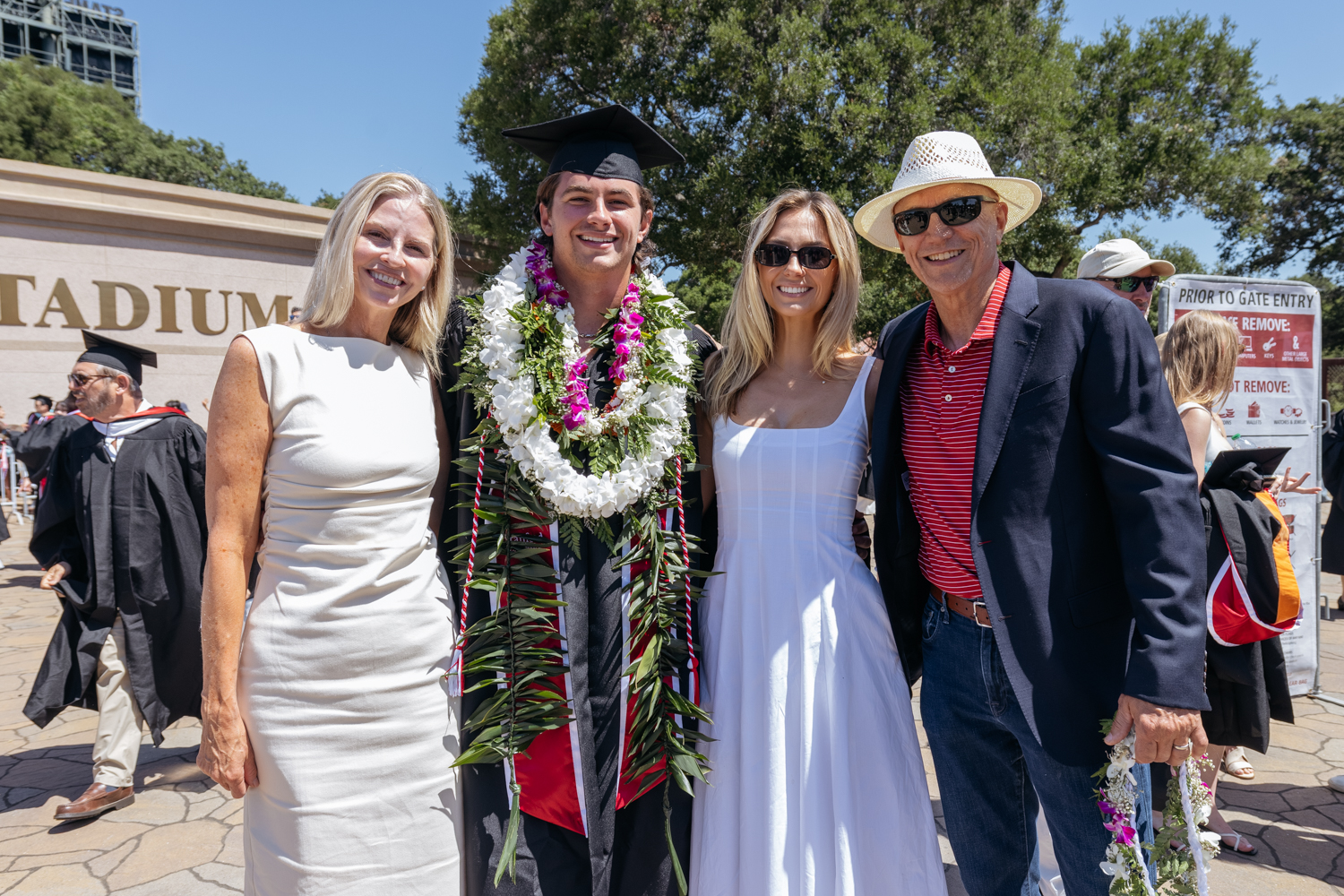 Graduate in cap, gown, and leis poses with family members outdoors after commencement ceremony.