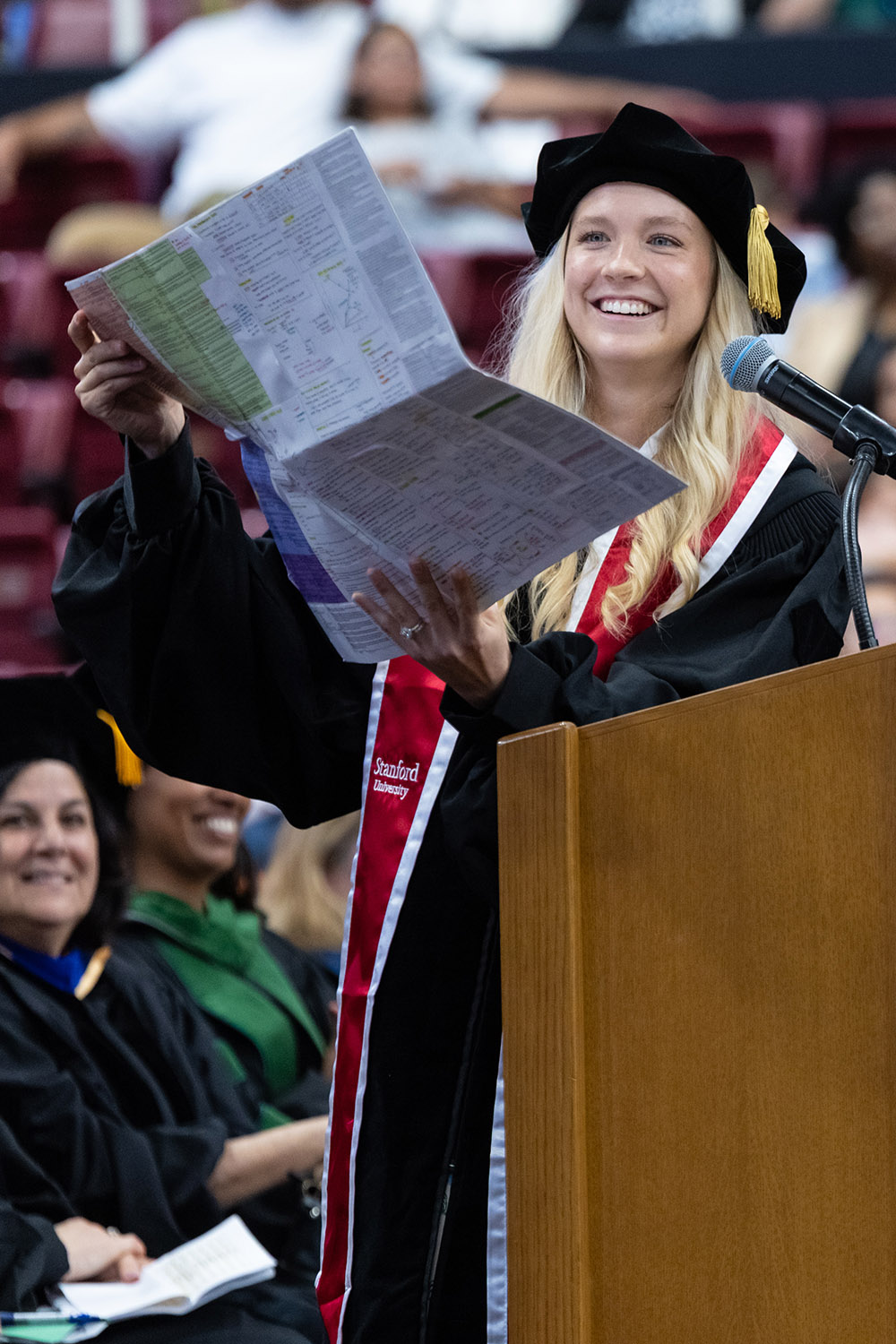 Smiling graduate at podium holds up large document during commencement speech.