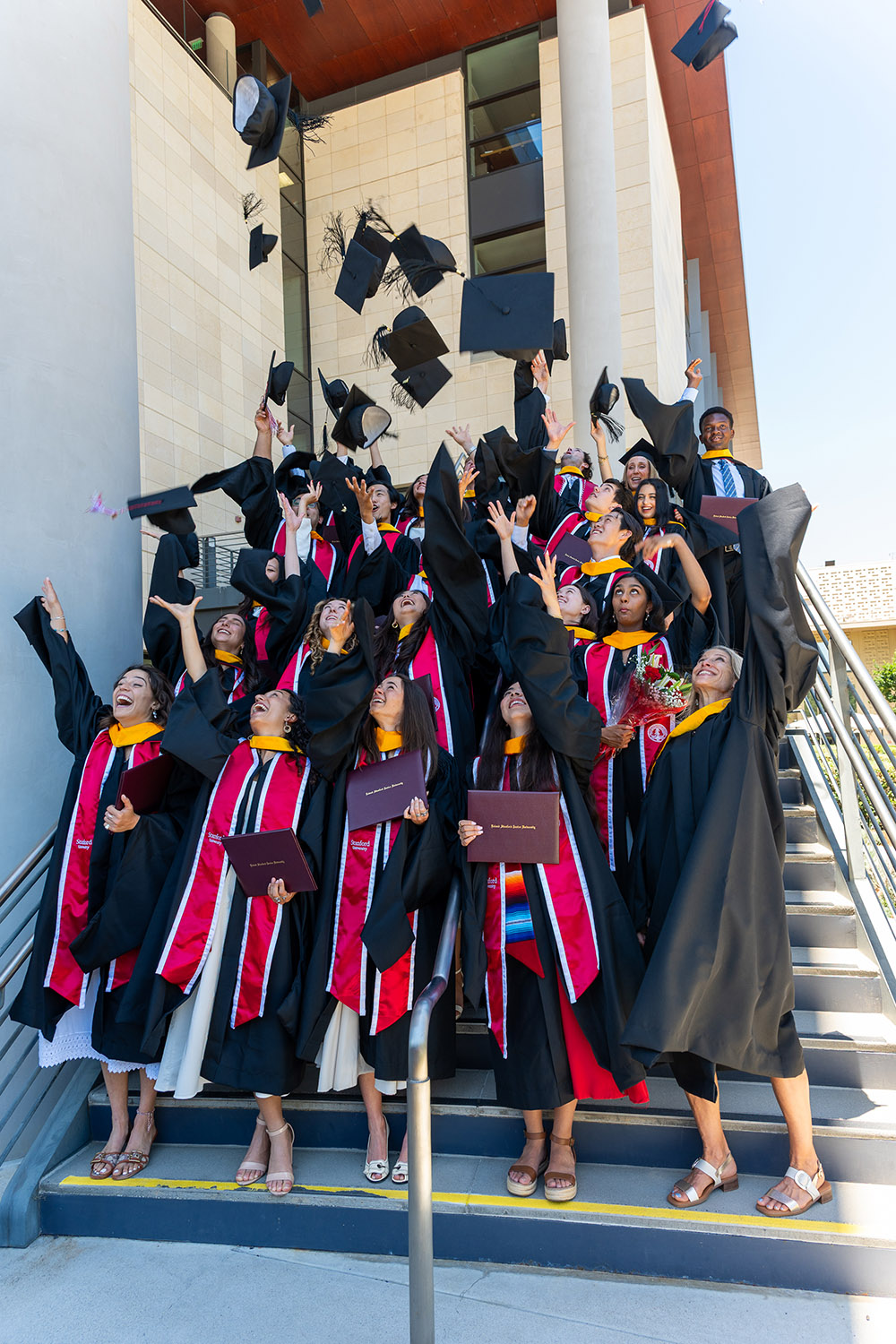Graduates celebrate by throwing their caps in the air while holding diplomas, gathered on a staircase following their graduation ceremony.