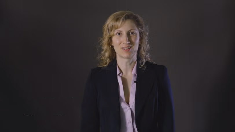 Pamela Schleissner, a woman with curly hair stands in front of a dark background, wearing a blazer and blouse, representing a research role.