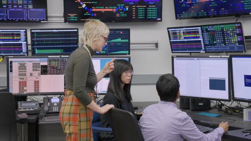 A team of three people discuss data at a control desk with multiple screens displaying information and graphs.