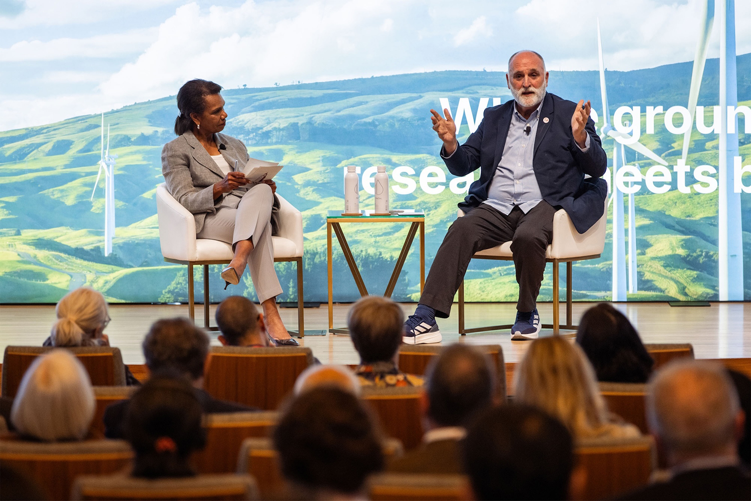 A panel discussion featuring Condoleezza Rice and Jose Andres on stage with a green landscape backdrop and wind turbines, engaging an audience.