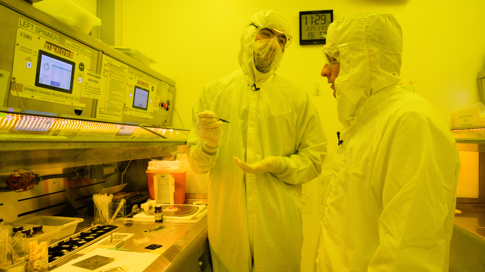 Two people in protective gear discuss procedures near specialized lab equipment, with various tools and documents visible on the table.