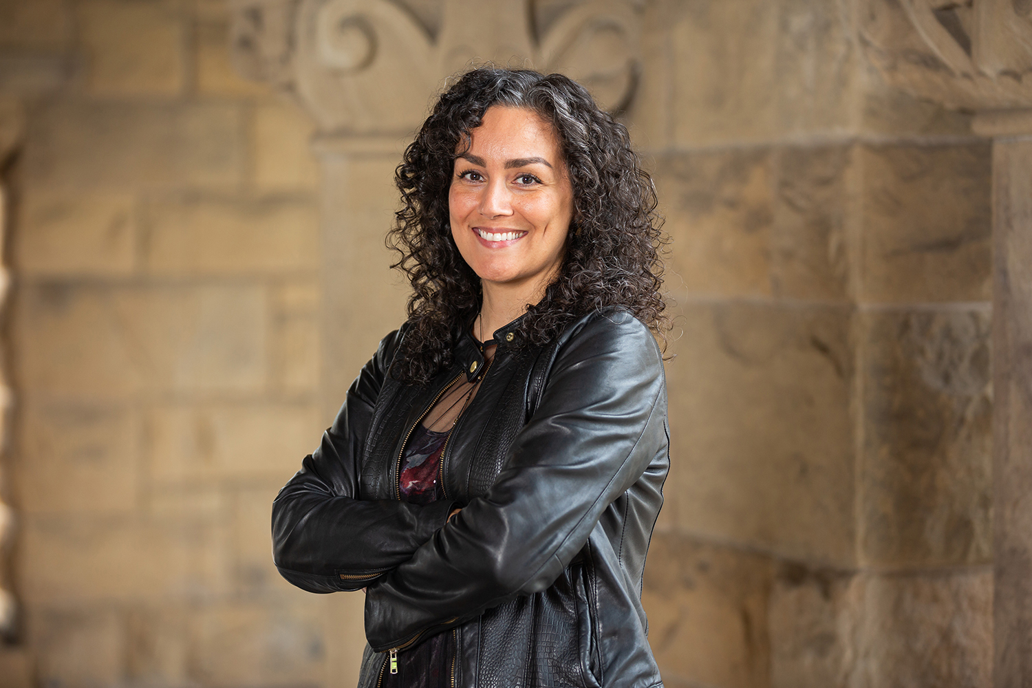 Rose Salseda posing and smiling for a profile photo and wearing a black leather jacket while standing with folded arms before a stone wall.