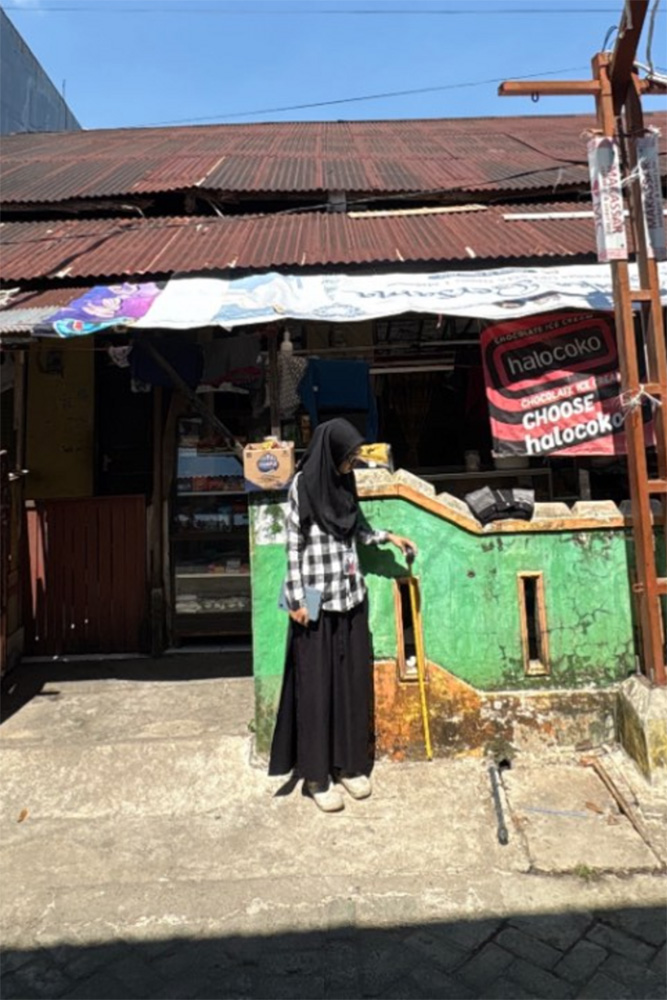 A woman in a hijab stands at the counter of a small neighborhood shop with a rusted tin roof.