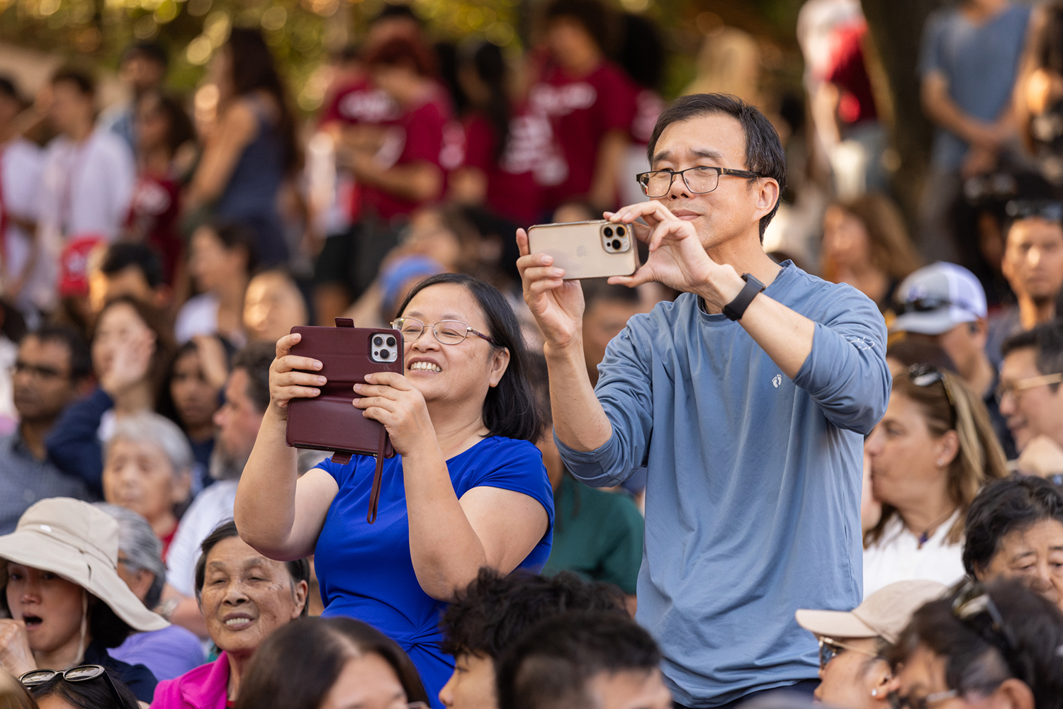 Two attendees hold up smartphones to photograph the ceremony from their seats in the crowd.