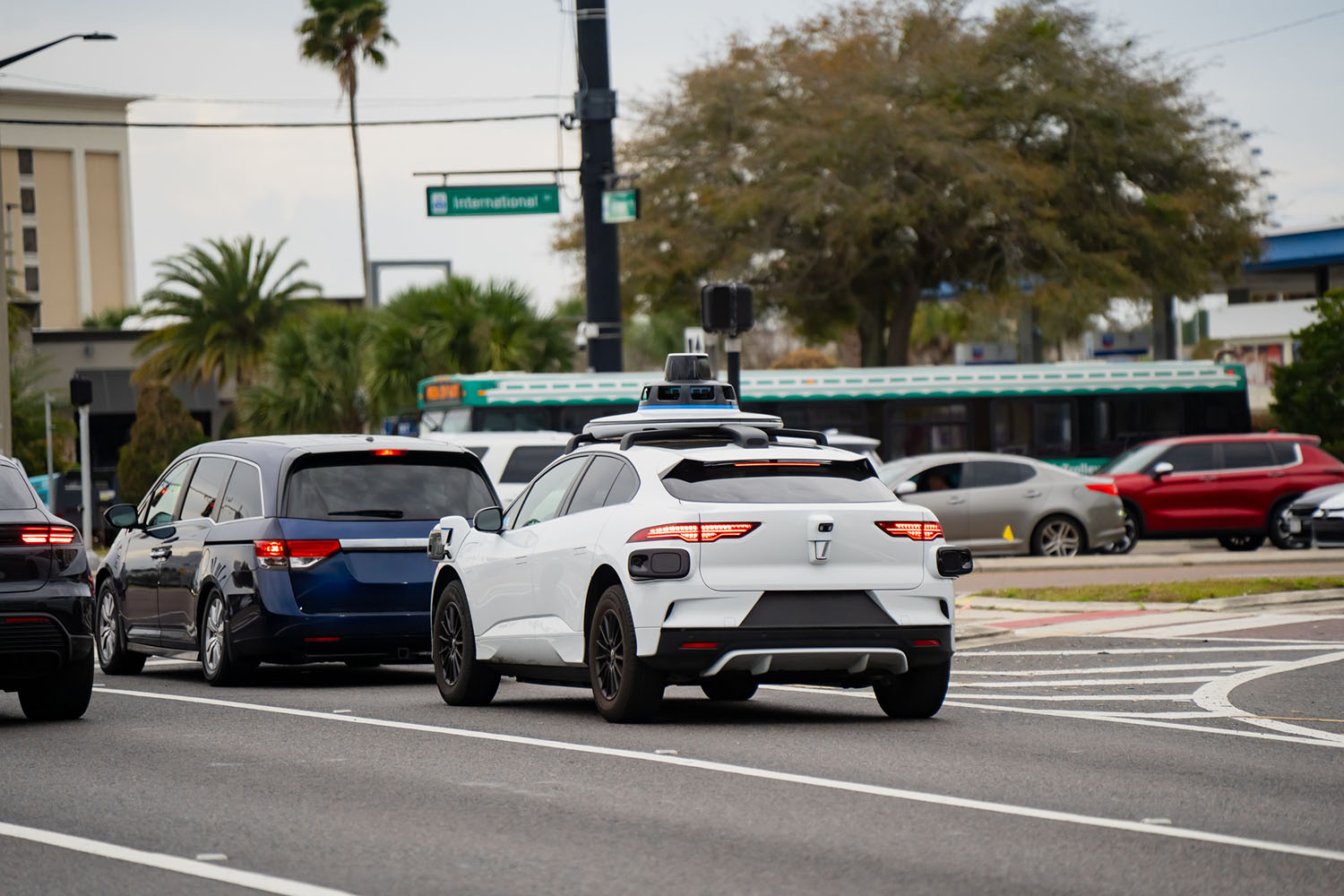 White self-driving car with roof-mounted sensors waits in traffic at a busy intersection.