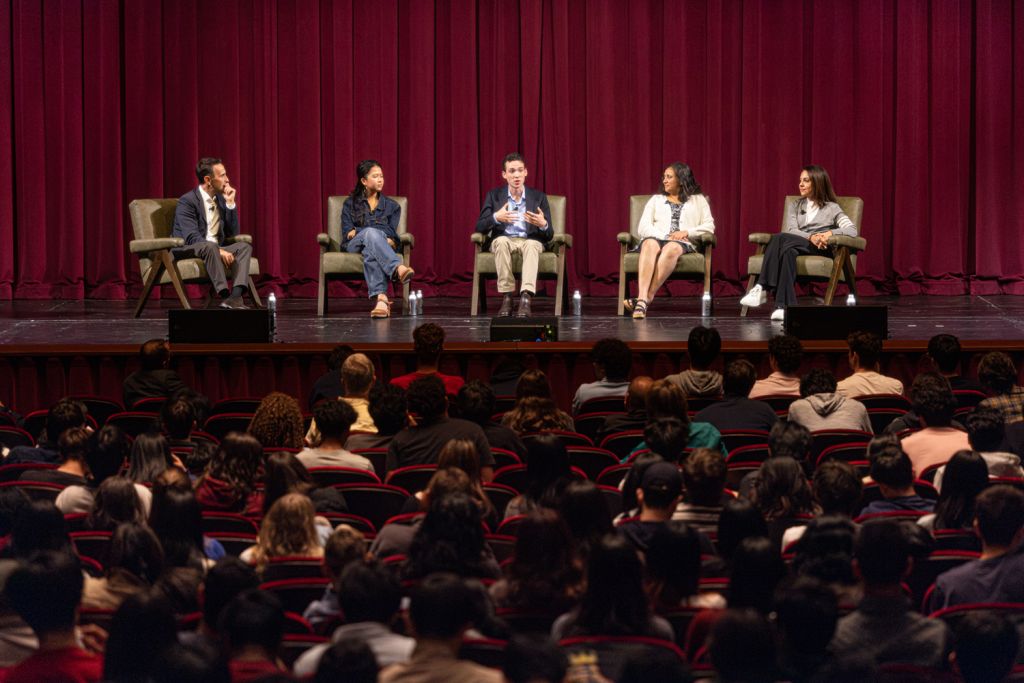 Five panelists sit on a stage before a red curtain, facing a large student audience in a theater.