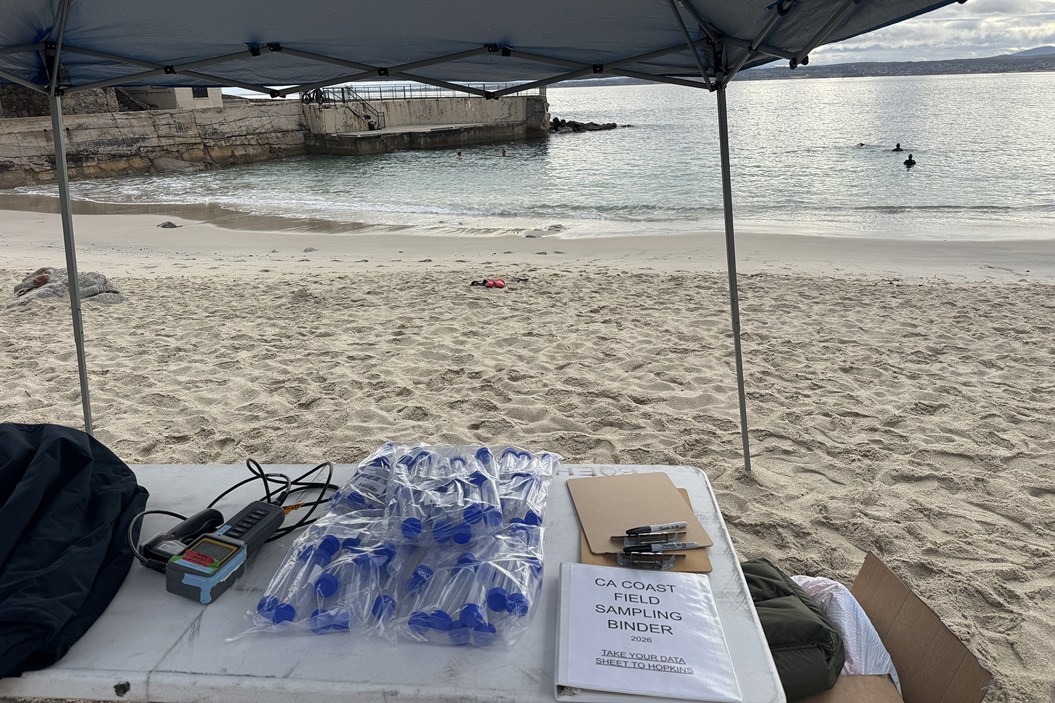 A field table under a canopy holds a water meter, sample tubes and a sampling binder, with the beach beyond.