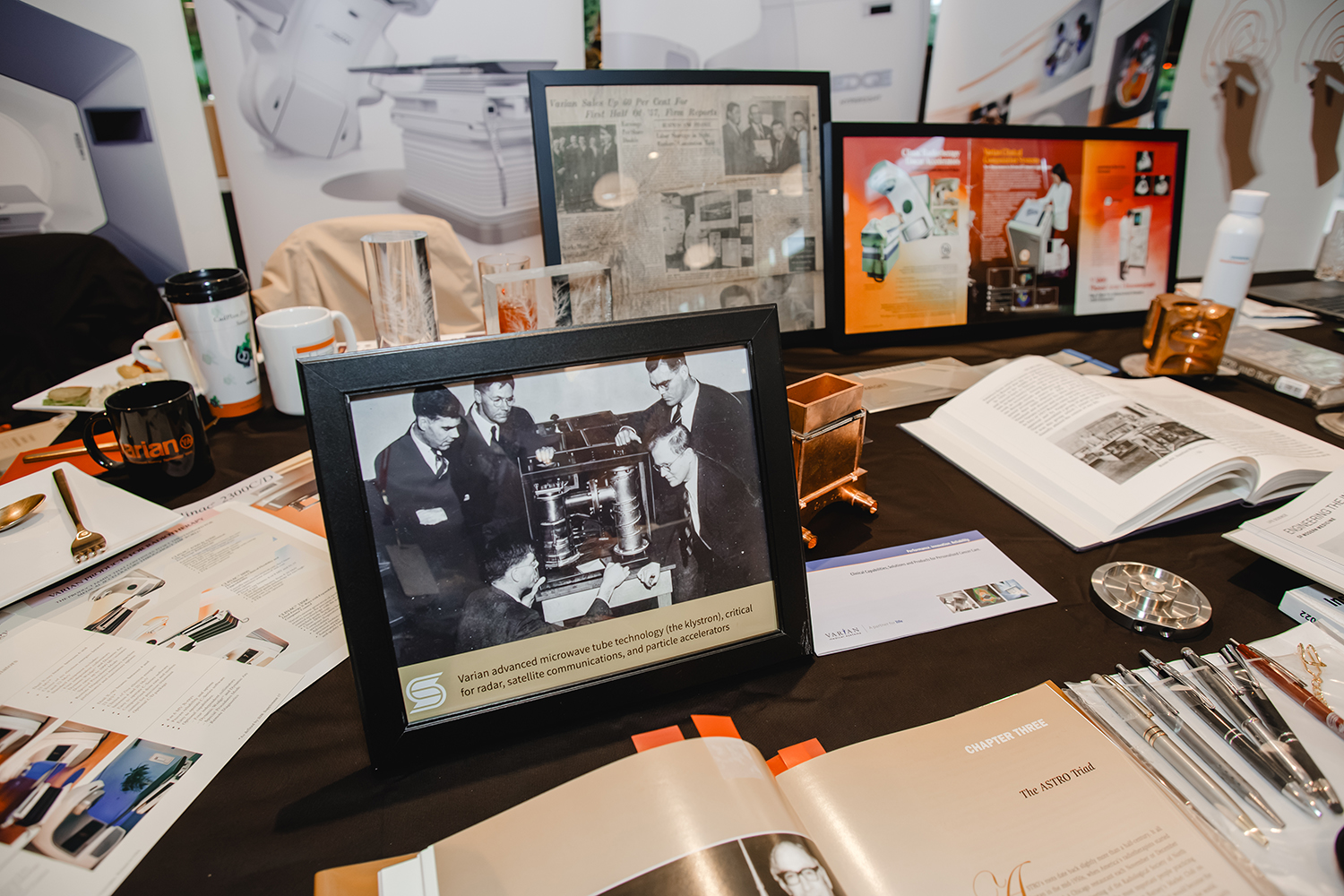 Varian exhibit table with a framed historic photo of Varian brothers and others examining a klystron, plus vintage clippings, books, and brochures.