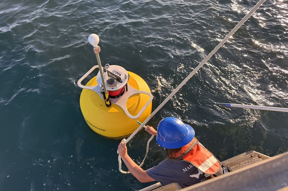Worker in a hard hat and life vest leans from a boat to secure a line to a yellow research buoy equipped with sensors.
