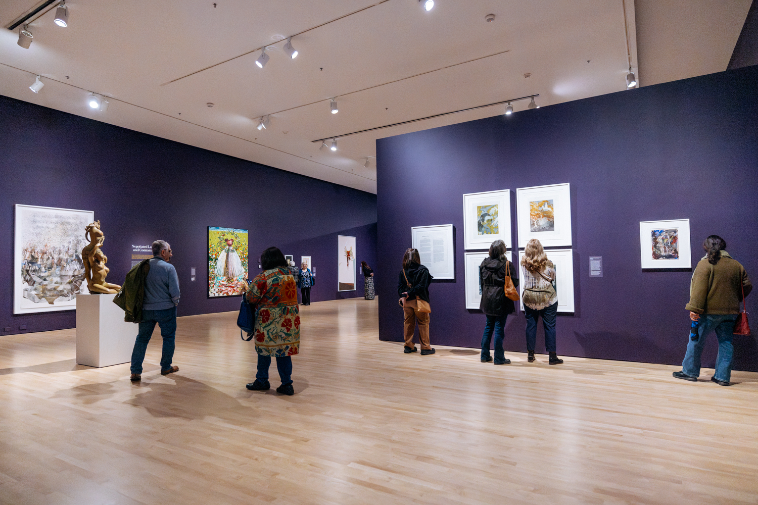 A deep purple gallery space features visitors engaging with a variety of artworks, including a bronze sculpture displayed on a pedestal.
