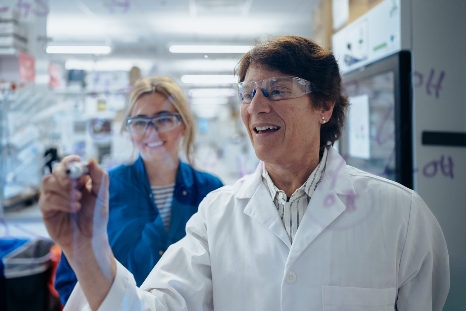 Carolyn Bertozzi writes notes on glass wall in her lab while another woman looks on.