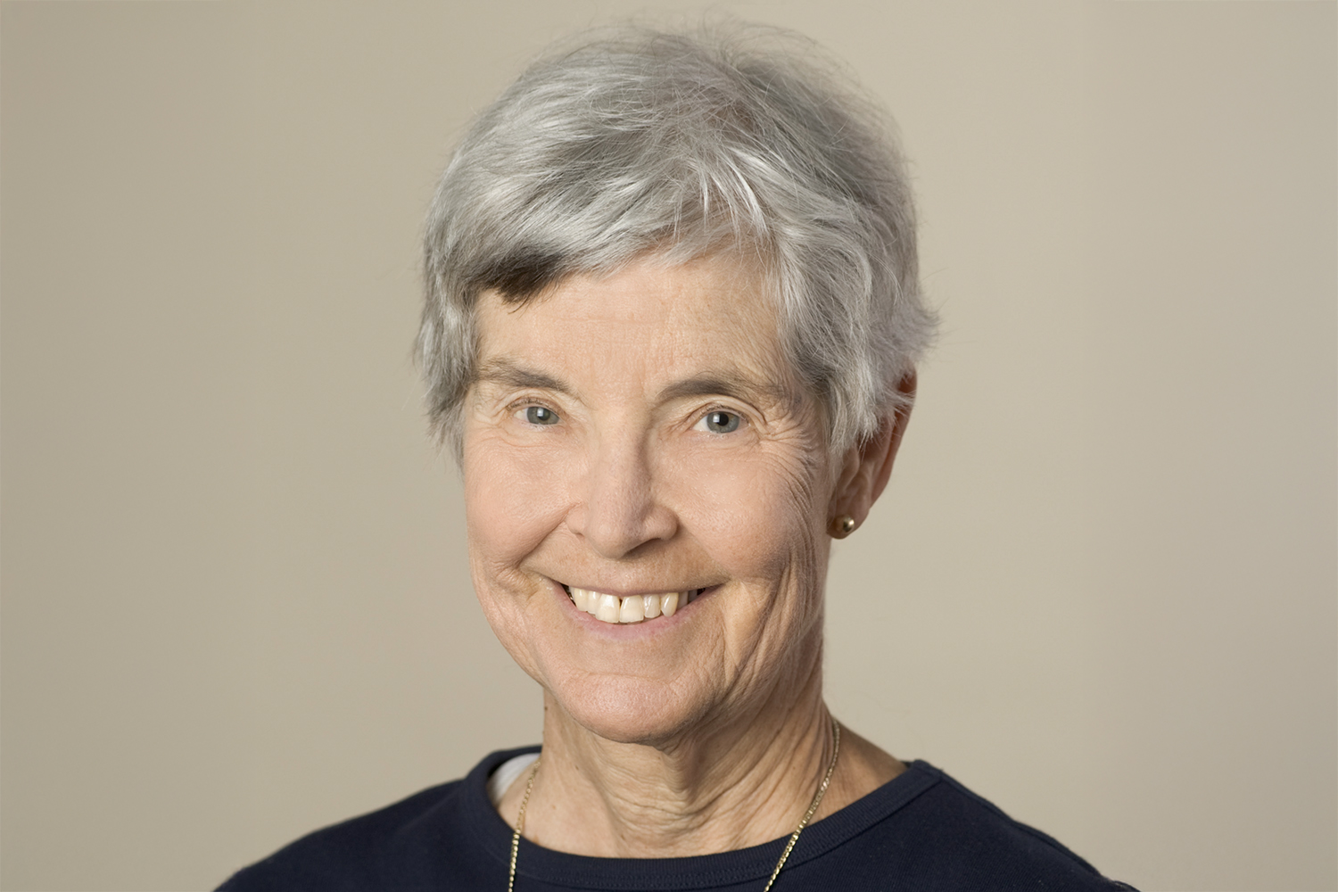 Close-up portrait of Alice Whittemore with short gray hair, wearing a dark shirt and a delicate gold necklace against a beige background.
