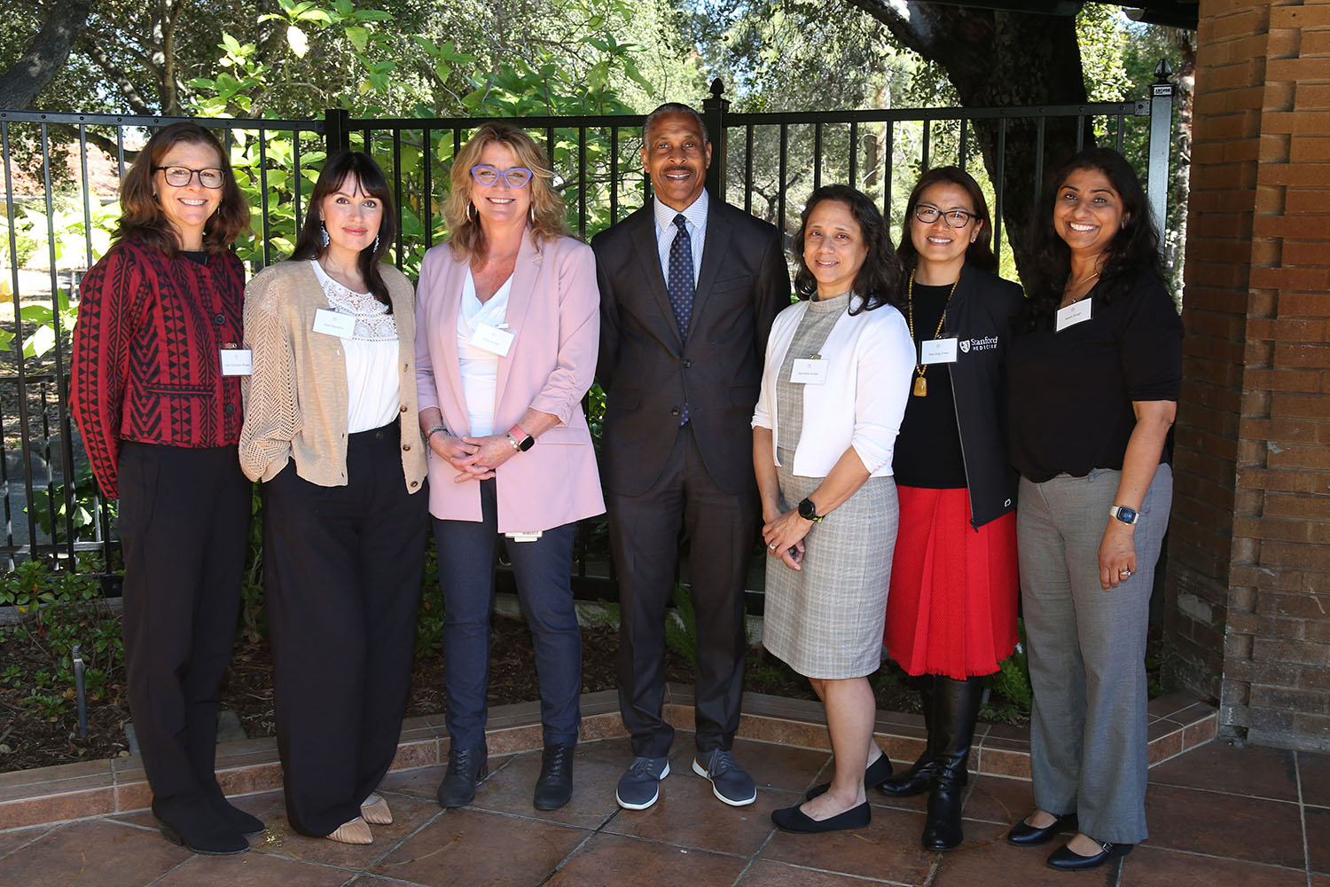 Group of seven people in business attire posing outdoors by a brick building and metal fence.