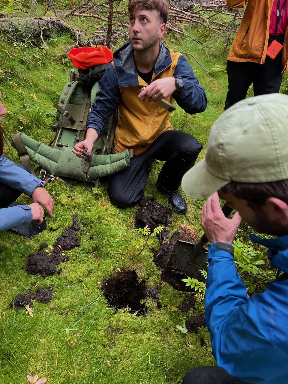 A group of people kneels in a green forest, examining soil samples from several dug holes while discussing their findings.