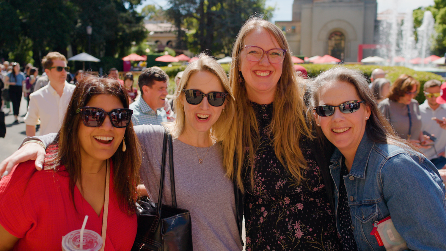 Open video Four people pose happily together at an outdoor event, with a fountain and numerous people in the background. in a modal