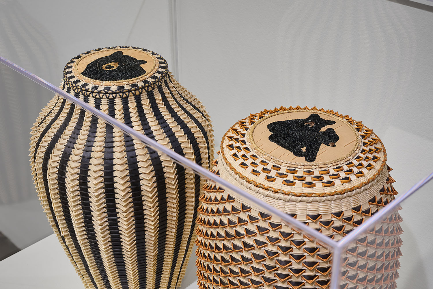 Two handwoven baskets with geometric patterns and designs of bears on the lids displayed in a glass case.