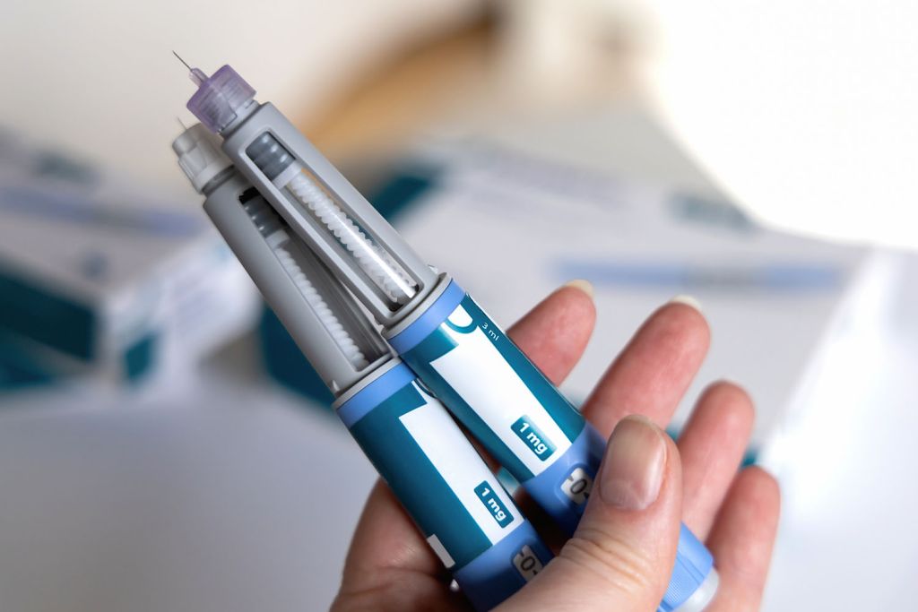 Hand holds two insulin pens with needle attached, with blurred packaging in the background.