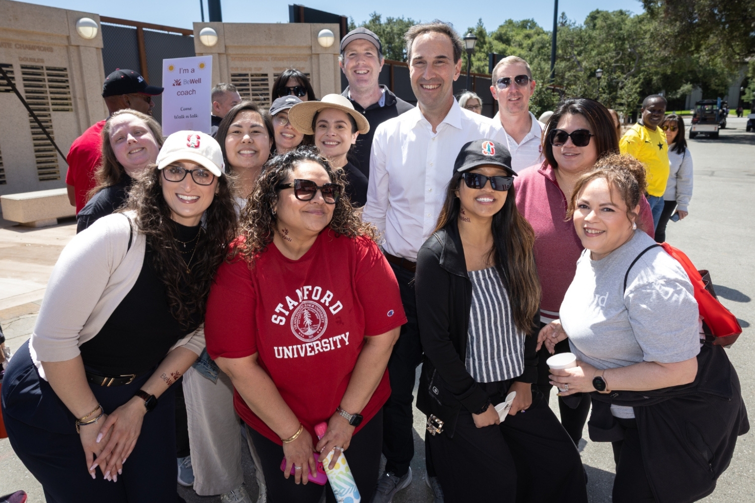 A group of people posing together, smiling and wearing Stanford apparel, in front of a sign promoting wellness at an event.