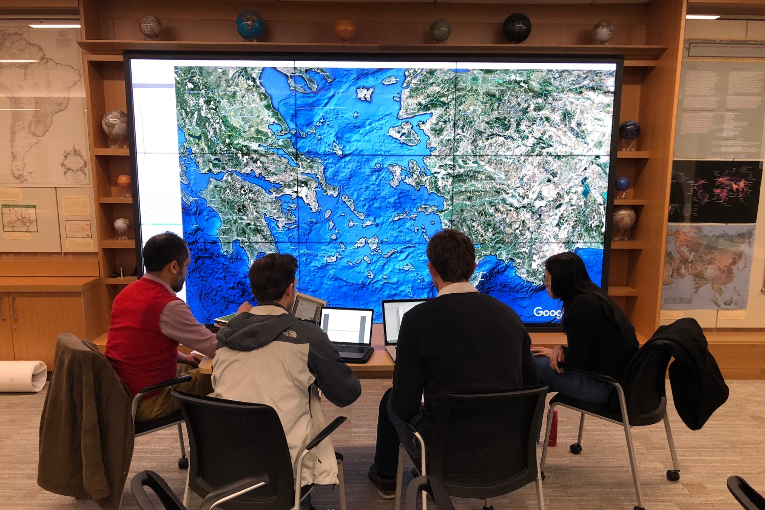 Four people view a large digital map on a wall screen while working on laptops in a collaborative setting.