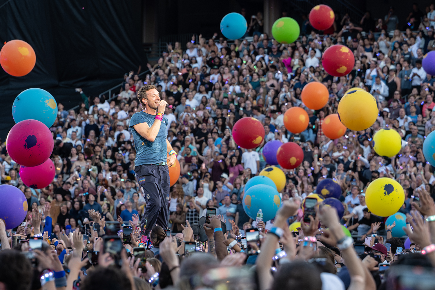 Coldplay performing on stage as colorful beach balls float above a cheering crowd, with many fans recording the moment on their phones.