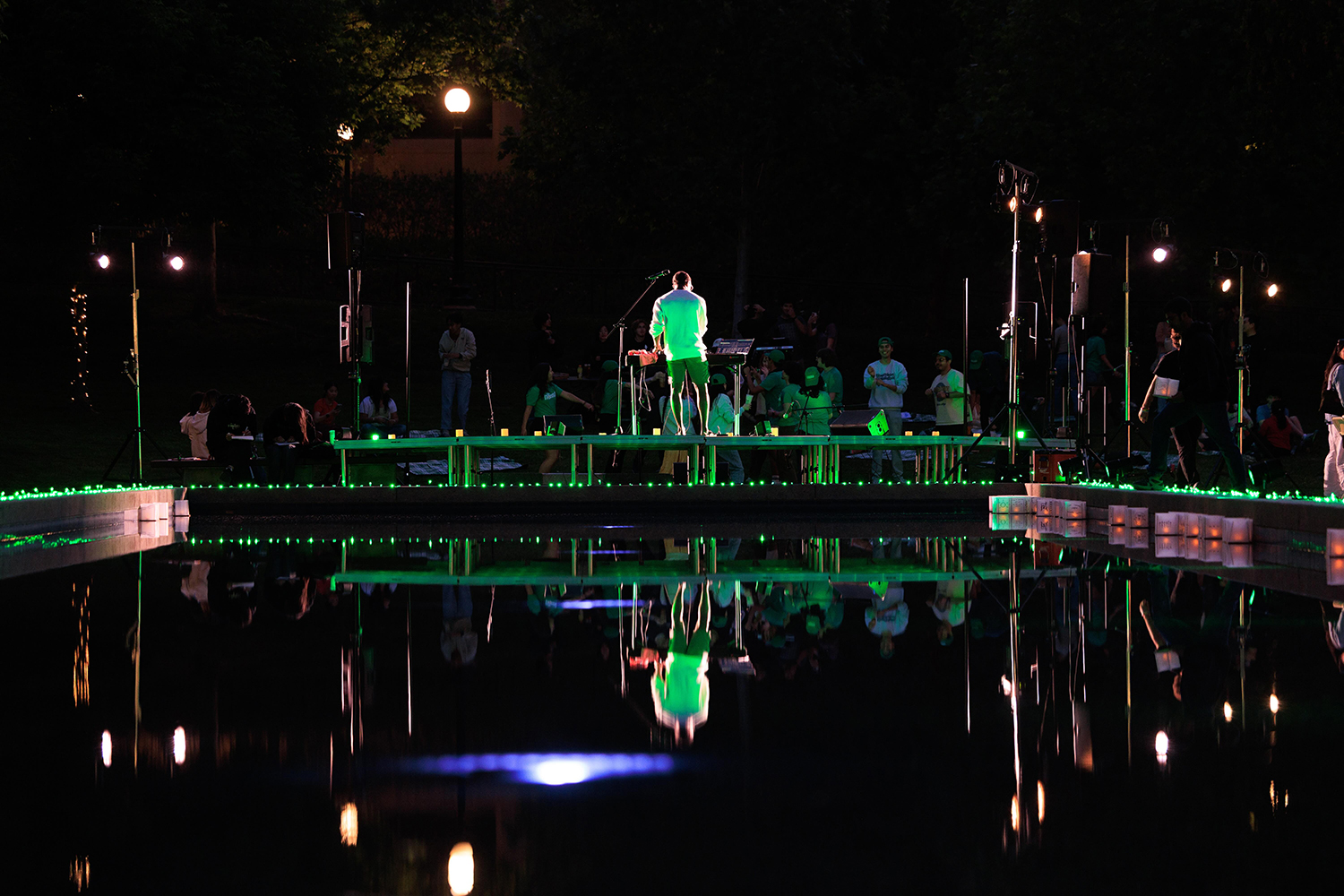 DJ stands on a stage lit with green lights, overlooking a crowd by Terman pond at the evening event.