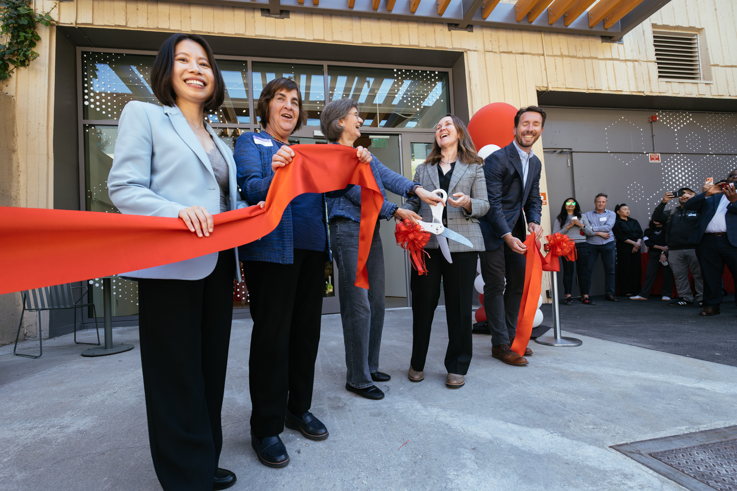 A group of people holds a large red ribbon, preparing to cut it at a ribbon-cutting ceremony, surrounded by onlookers.