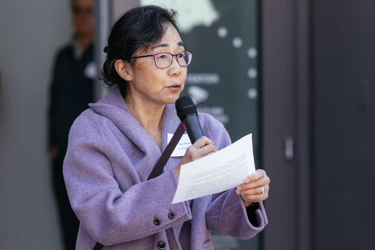 A speaker holds a microphone and notes while addressing an audience during an outdoor event.