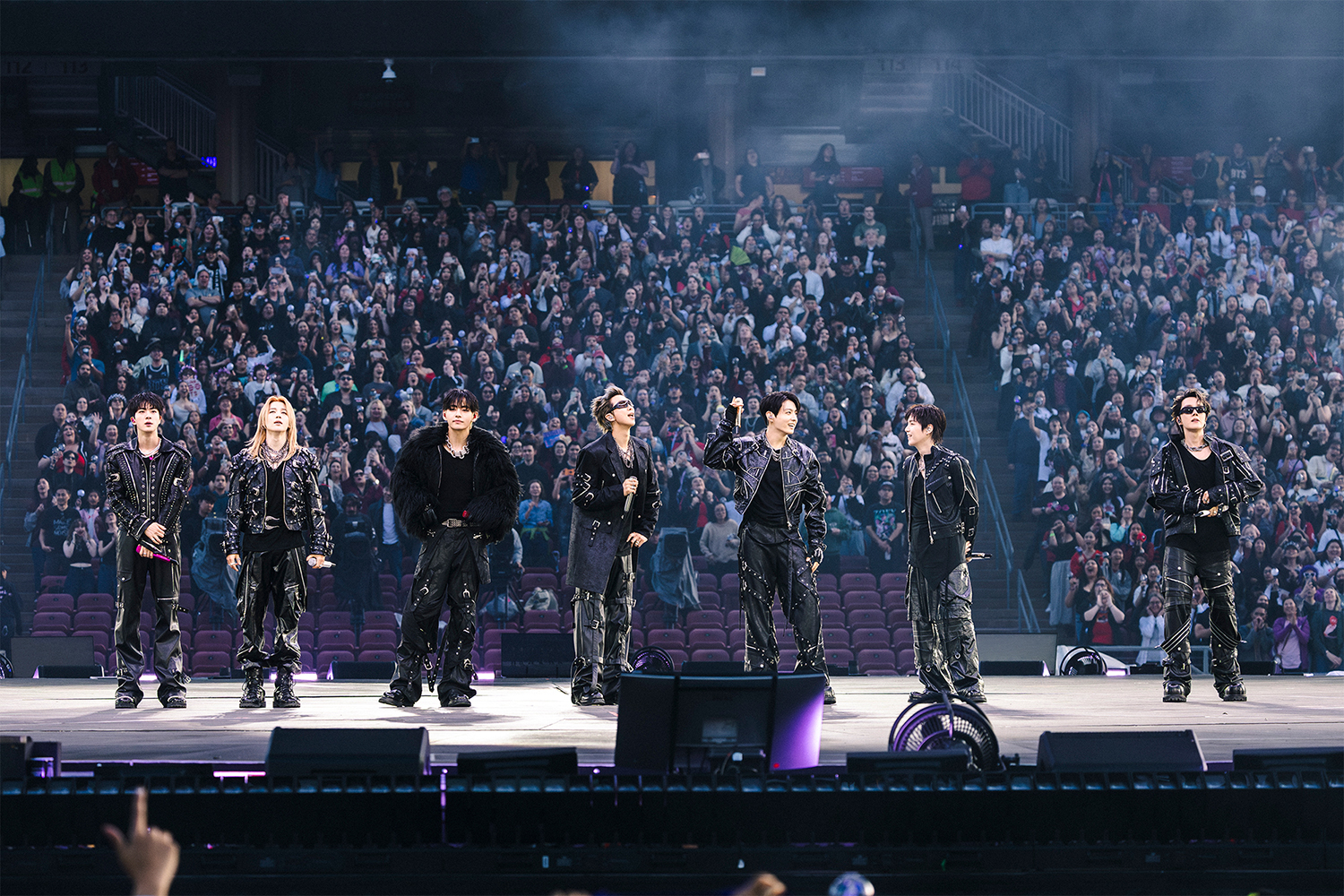 BTS performs on stage in black outfits, surrounded by an enthusiastic audience in a large arena.
