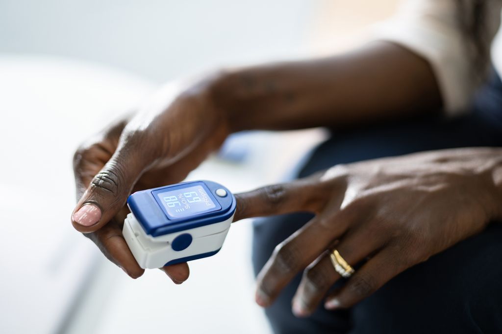 A person measures their oxygen levels using a finger pulse oximeter, displaying readings on a digital screen.