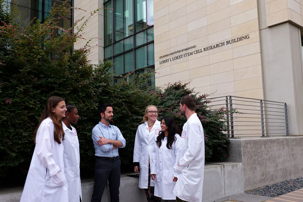 A group of five researchers in lab coats stand outside the Lorry I. Lokey Stem Cell Research Building, engaging in conversation and smiling.